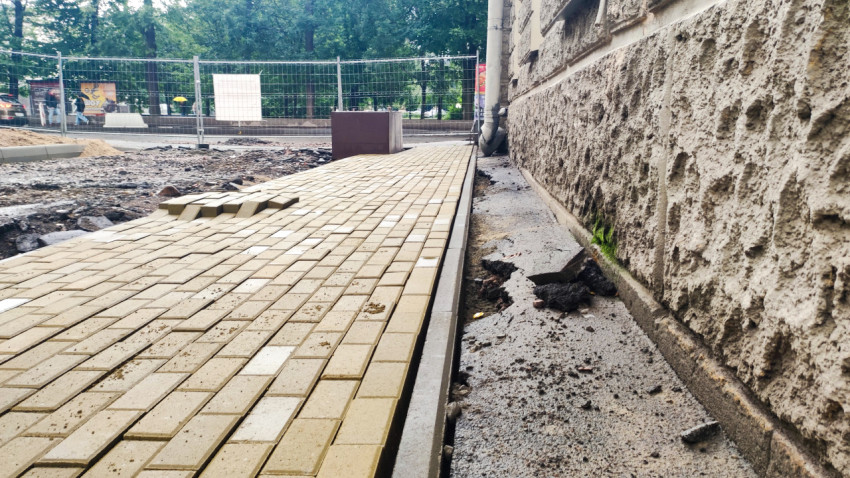 Неровно уложенная тротуарная плитка вдоль фасада и разрушенный асфальт у стены здания