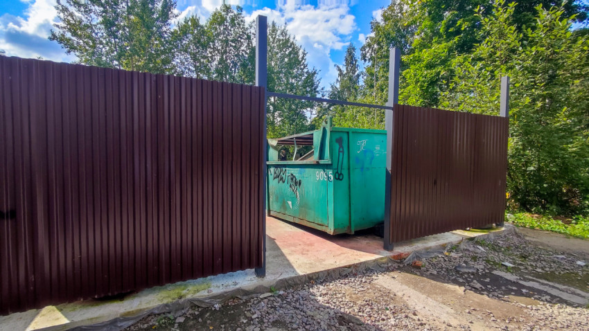 Въезд на ограждённую контейнерную площадку с зелёным мусорным контейнером и граффити