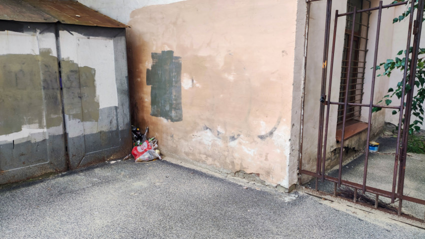 Угол двора с облупленной стеной, металлическими воротами и пакетом мусора у хозяйственной постройки