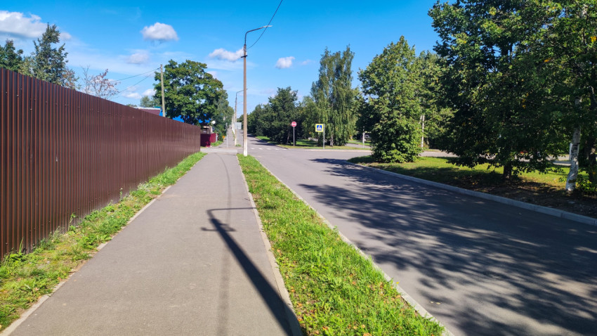 Тротуар вдоль коричневого забора и пустая дорога с тенями деревьев у перекрёстка
