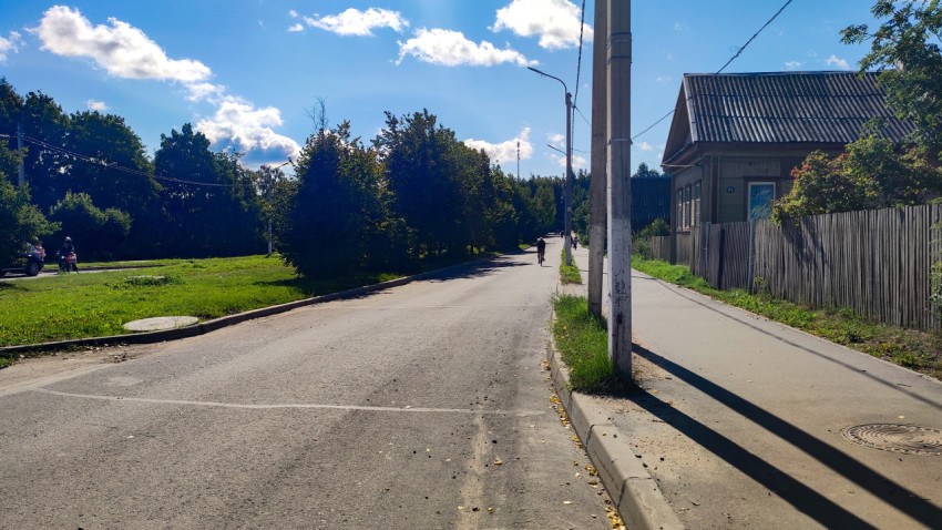 Улица частного сектора с тротуаром, деревянным домом и высоким столбом освещения на фоне зелёных деревьев
