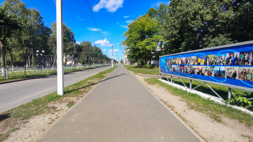 Длинный тротуар вдоль дороги и большой синий баннер на ограждении у зелёной зоны