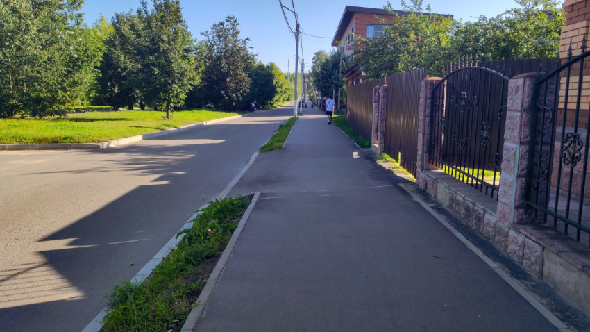 Длинный тротуар вдоль дороги рядом с забором, газоном и деревьями частного сектора