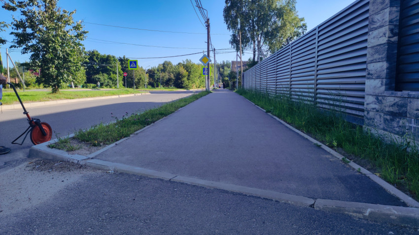 У перекрёстка стоят дорожные знаки, а вдоль забора тянется широкий тротуар в тени деревьев