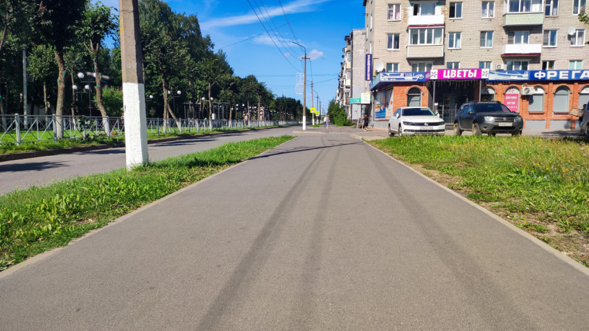 Ровный тротуар вдоль дороги, припаркованные машины и магазинные вывески на первом этаже дома