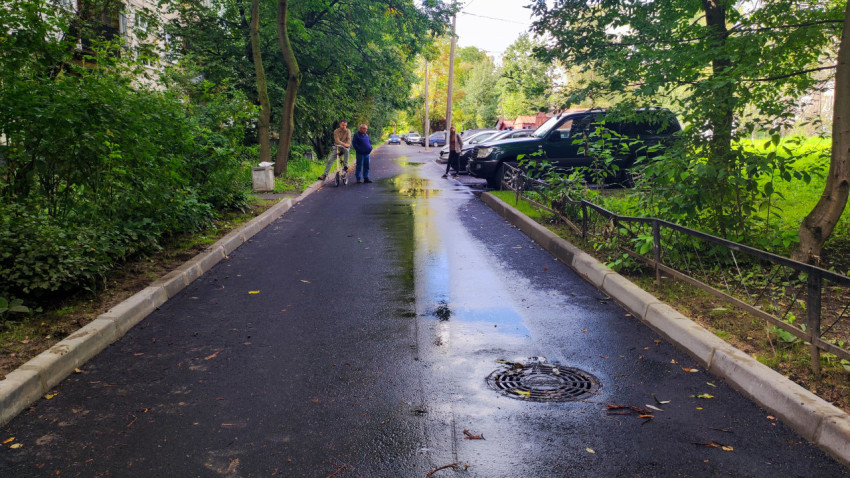 Мокрый дворовый проезд с лужами, люком и припаркованными машинами у жилых домов