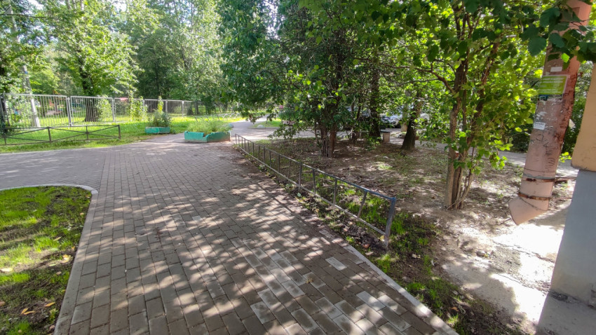 Дворовая дорожка из плитки идёт между деревьями и домом, рядом стоят зелёные клумбы