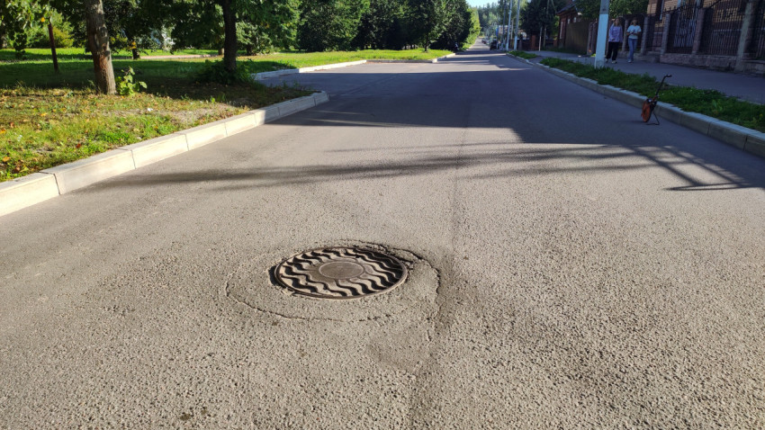 Люк на свежем асфальте посреди тихой улицы с бордюрами и тенями от деревьев