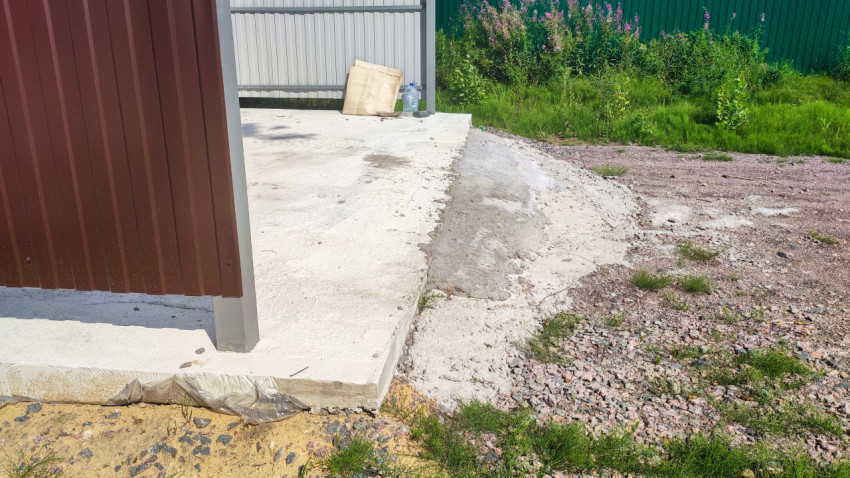 Бетонная площадка контейнерной зоны и коричневое ограждение с гравийной отсыпкой и травой