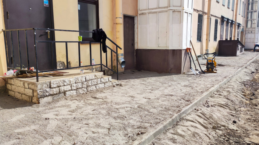 Подготовка песчаного основания под тротуар у входа в здание с инструментами и виброплитой