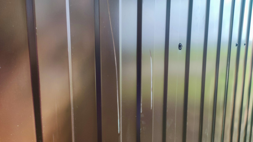 Царапины и вмятина на коричневом профлисте ограждения контейнерной площадки крупным планом