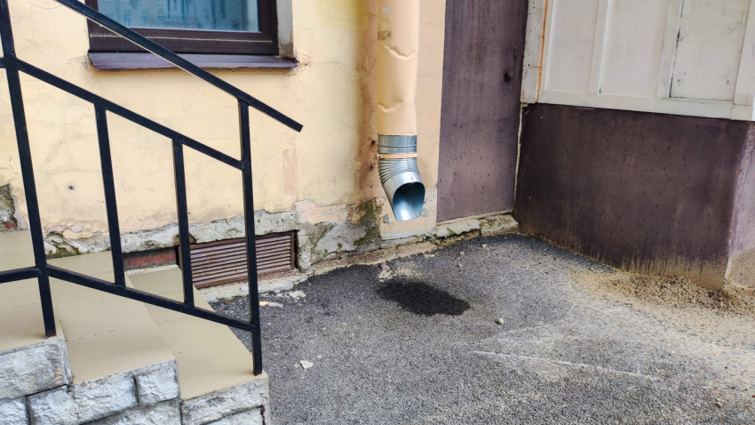 Водосточная труба у фасада дома и мокрое пятно на дворовом покрытии возле основания стены