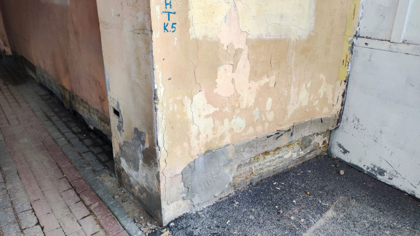 Угол здания с облупленной штукатуркой, ремонтными заплатами и синей маркировкой у тротуарной плитки