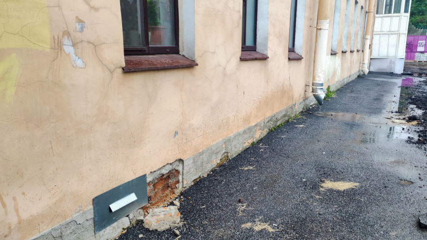 Повреждённый цоколь жилого дома с трещинами штукатурки и лужами у водосточной трубы