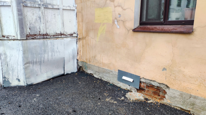 Ржавые металлические створки и следы намокания на углу здания с повреждённой отделкой цоколя