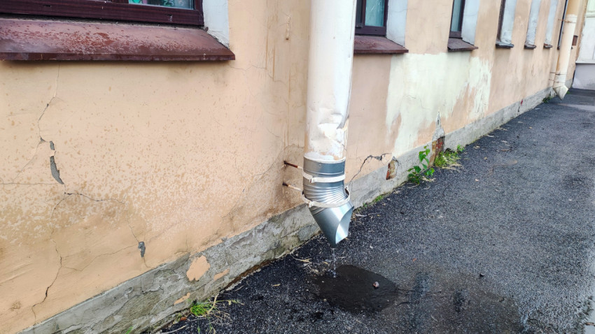 Нижний выход водосточной трубы с коленом и лужей у повреждённого цоколя здания