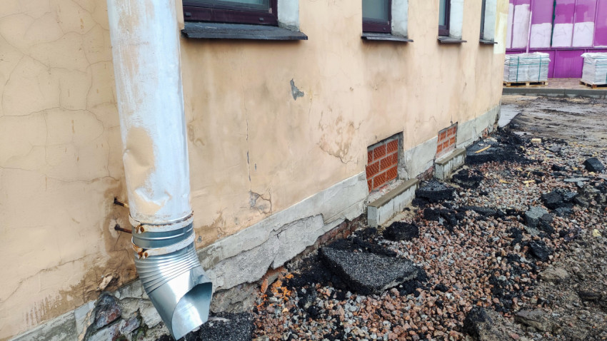 Водосточная труба с гибкой гофрой и металлическим отводом на потрескавшемся фасаде у цоколя