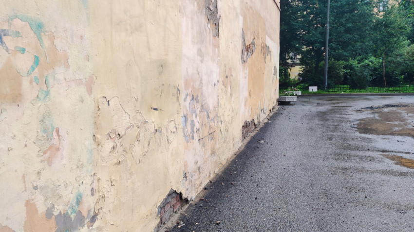 Длинная стена здания с трещинами и пятнами на штукатурке и мокрым асфальтом с лужами у фасада