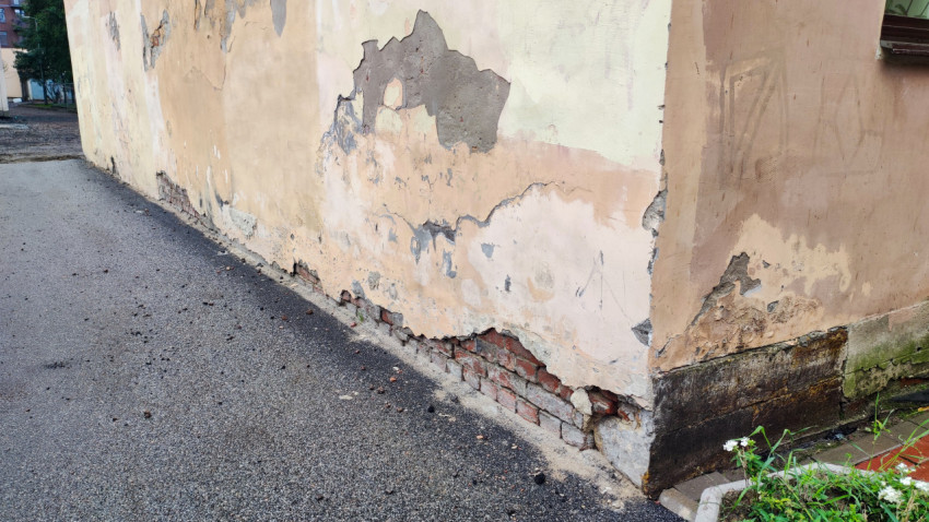 Угол здания с облупившейся штукатуркой, трещинами и зелёным налётом у основания цоколя