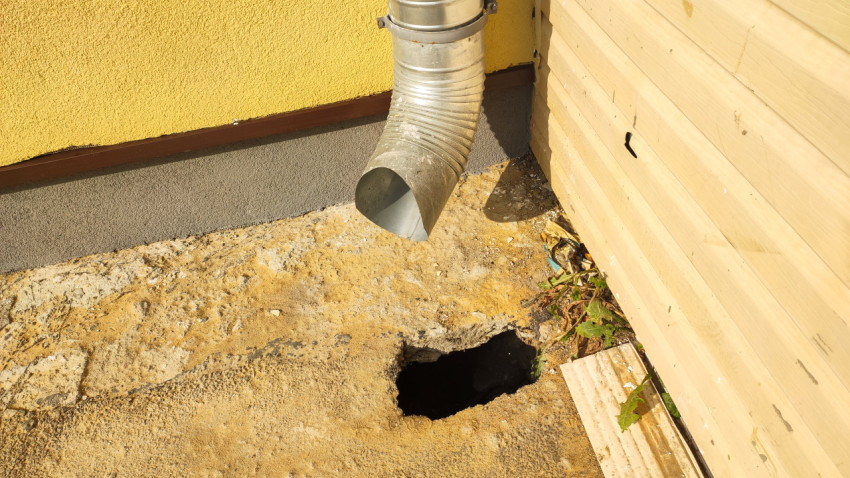 Водосточная труба с отводом выходит на бетон у стены дома рядом с провалом в грунте