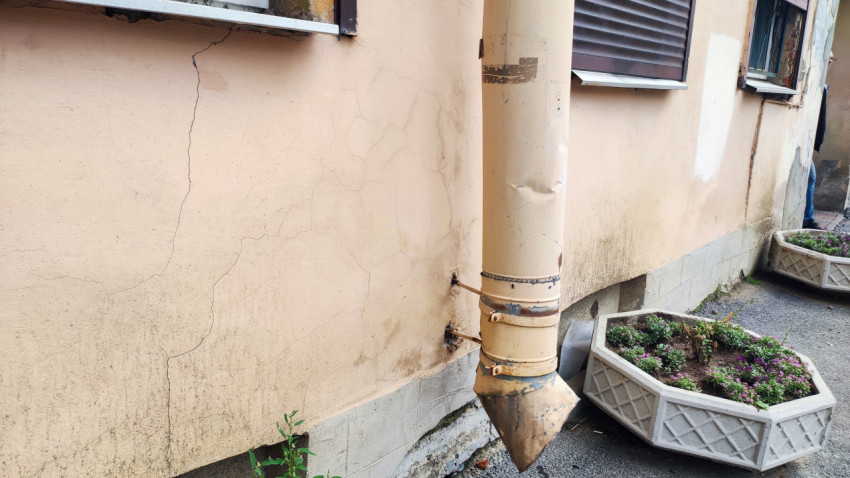Водосток у стены дома и повреждения отделки рядом с бетонным вазоном на тротуаре