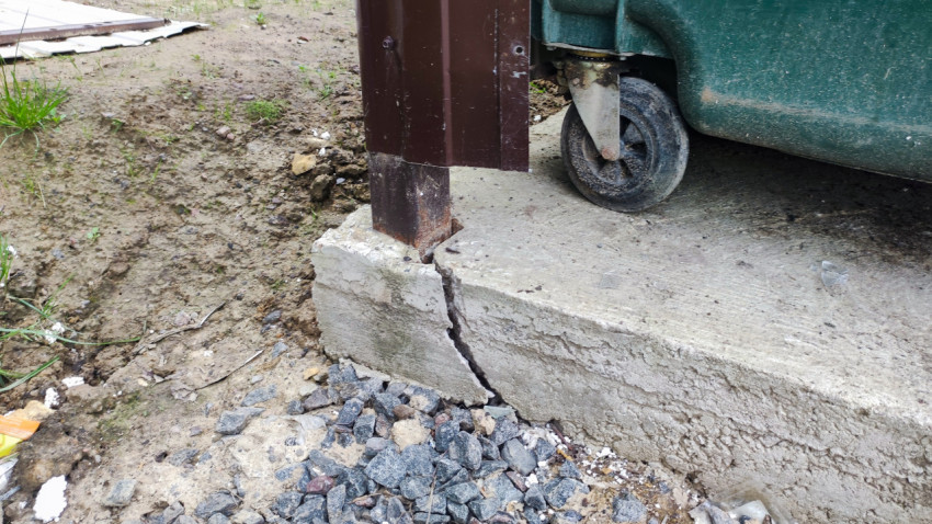 Трещины и сколы бетонного основания у металлической стойки рядом с мусорным контейнером