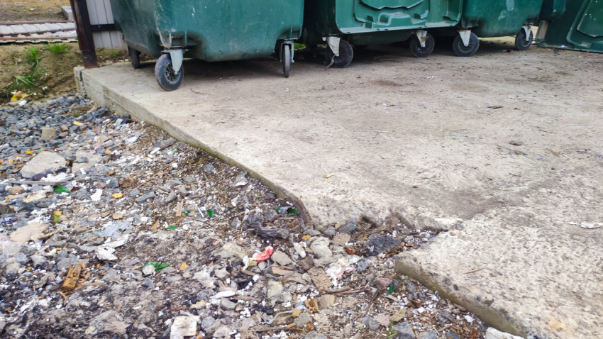 Мусор и каменная крошка у края бетонной площадки с колёсами контейнера