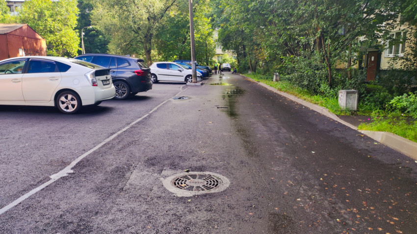 Свежий асфальт с мокрым пятном и люком на проезде между припаркованными автомобилями