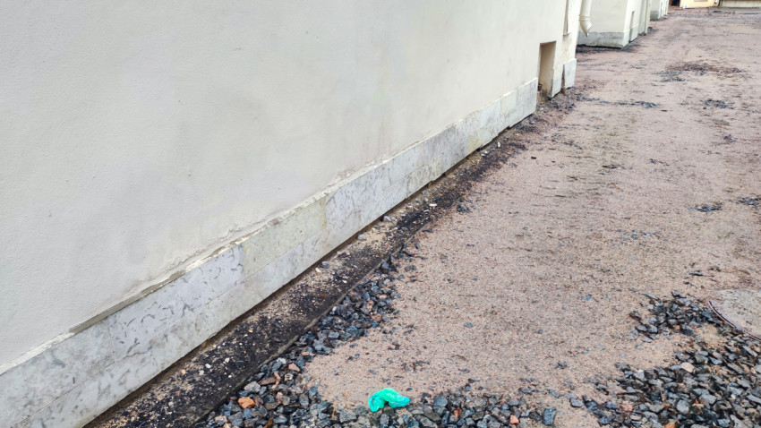 Протяжённый белый фасад без окон с серым цоколем и грунтово-гравийным покрытием вдоль стены