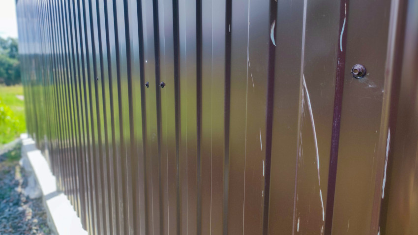 Металлический забор из профнастила на фоне поля и деревьев