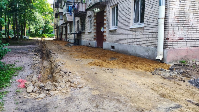 Разобранный участок дворовой дороги у кирпичного дома с песком, камнями и выемкой вдоль фасада