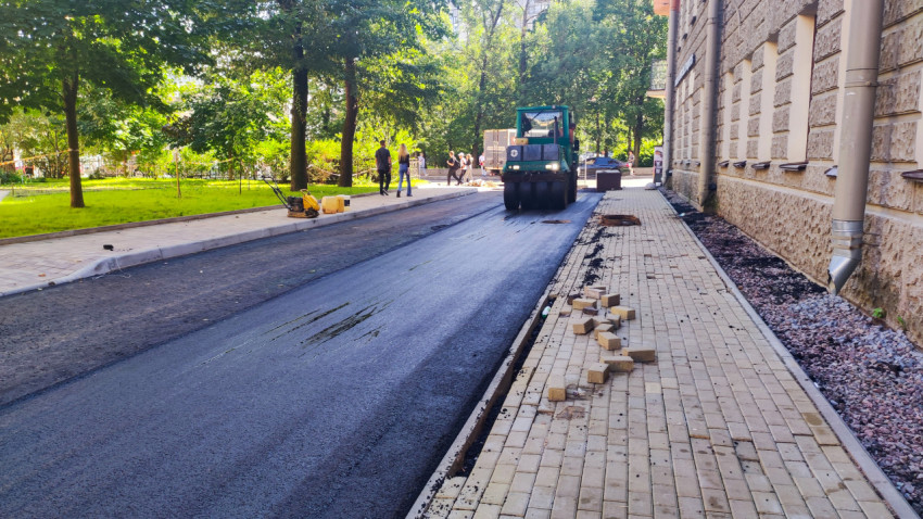 Каток уплотняет свежий асфальт на проезде рядом с тротуаром и рассыпанной плиткой