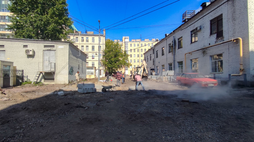 Рабочие и экскаватор на пыльной стройплощадке между кирпичными корпусами