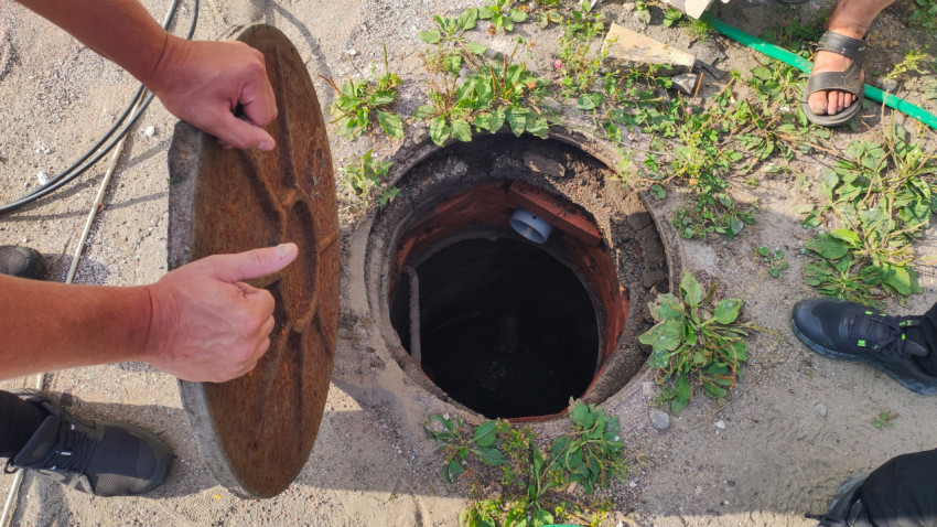 Человек приподнимает ржавую крышку люка, открывая шахту коммуникаций среди песка и травы