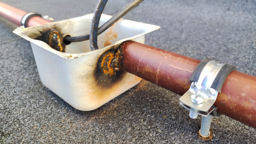 Крупный план металлической коробки на трубе с тёмными потемнениями и коррозией вокруг отверстий