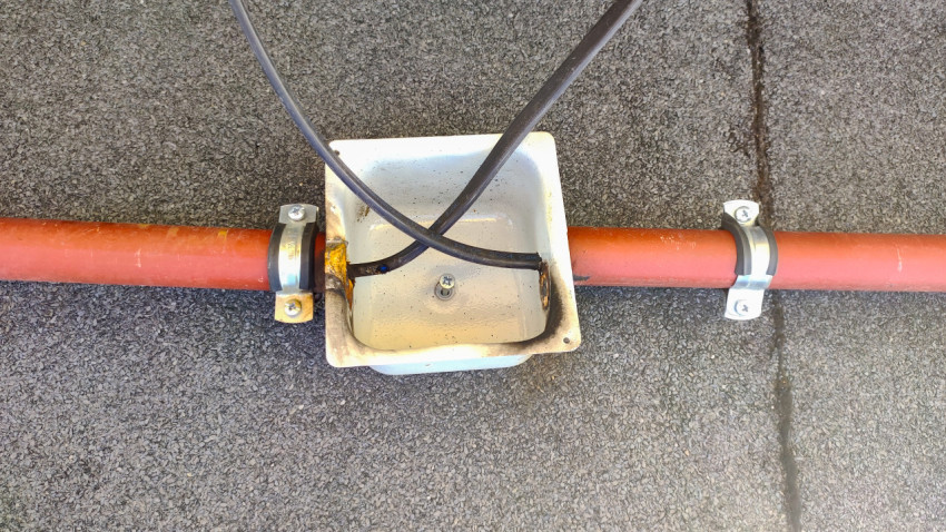 Вид сверху на металлическую коробку с кабелями, закреплённую на трубе хомутами на плоской кровле