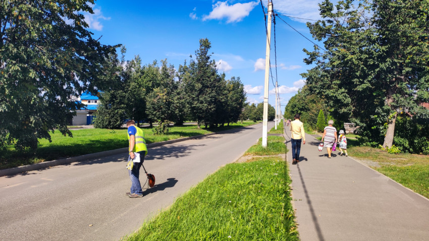 Рабочий катит измерительное колесо вдоль дороги, пока по тротуару идут прохожие