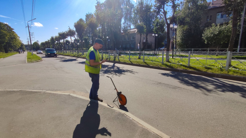 Мужчина в жилете стоит у края дороги с измерительным колесом и смотрит в телефон