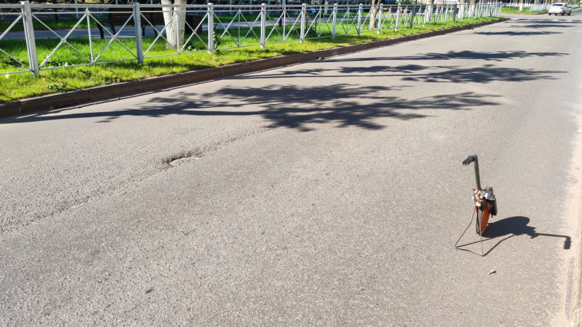 На пустой улице заметна выбоина, а рядом стоит измерительное колесо на ножке