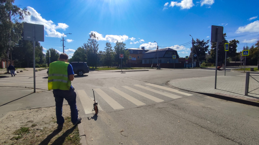 Рабочий в сигнальном жилете стоит у зебры на перекрёстке, рядом знаки и измерительное колесо