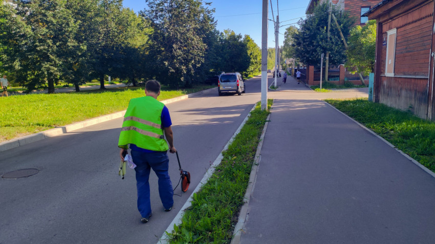 Рабочий с измерительным колесом идёт вдоль бордюра на улице у деревянных домов и деревьев