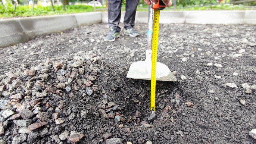 Замер глубины основания у бордюра рулеткой рядом с лопатой на дворовой дороге