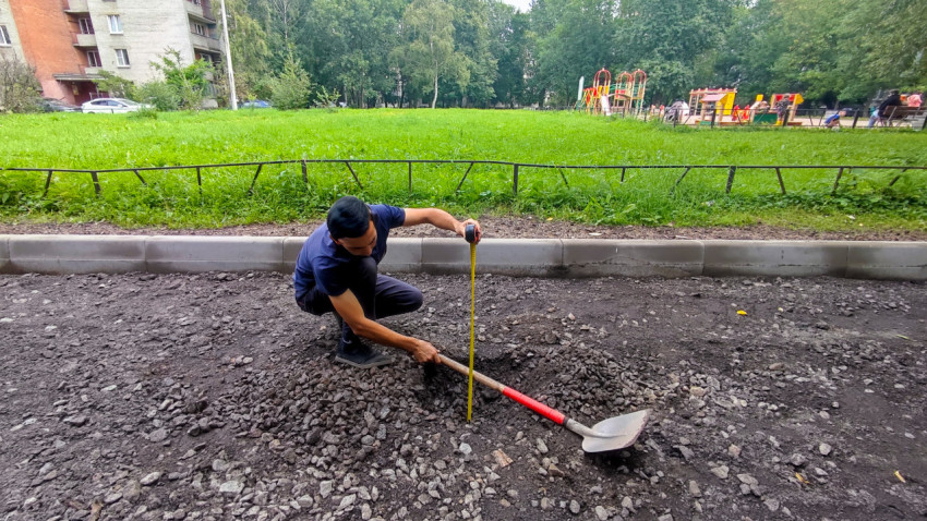 Рабочий измеряет глубину отсыпки у бордюра рулеткой на участке благоустройства двора