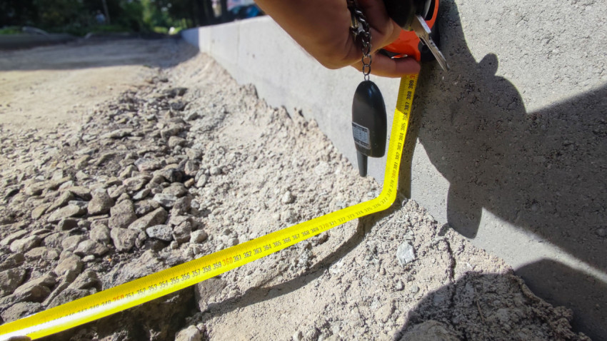 Замер глубины основания у бордюрного камня строительной рулеткой на участке дорожных работ