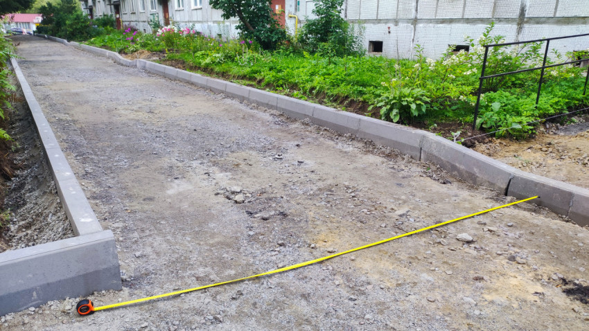 Рулетка, натянутая через щебёночный проезд у фасада жилого дома и клумбы