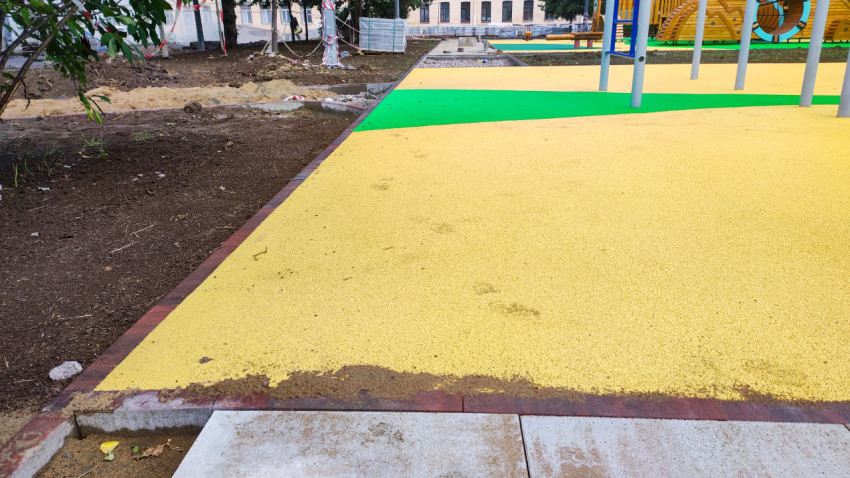 Желтое резиновое покрытие у кромки грунта и плитки на благоустроенной площадке