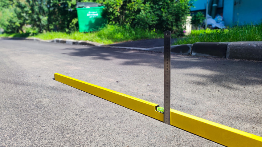 Строительный уровень на дороге и рейка у бордюра для замера уклона на дворовом проезде