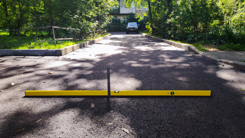 Контроль уклона дворовой дороги уровнем и линейкой на фоне бордюров и зелени