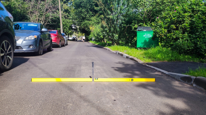 Измерение перепада асфальта уровнем и рейкой на дворовой дороге рядом с припаркованными машинами