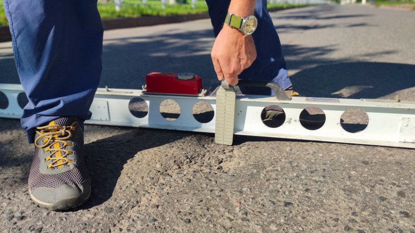 Рука с клин-промерником снимает показания под рейкой на зернистом асфальте